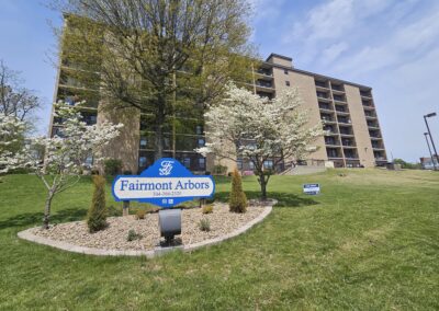 A view of a large eight story apartment building across a lush green lawn. a small rock garden with several trees is in front of the building and includes a sign that reads "Fairmont Arbors" with the phone number 304-366-2320