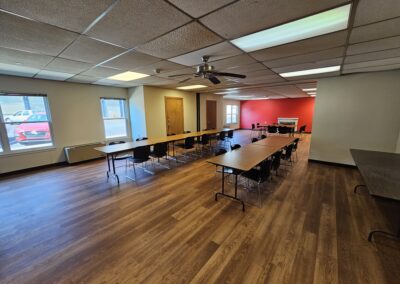 A community center space in an apartment building. There are two long tables in the middle of the room with chairs on either side and one shorter table on the far side of the room with eight chairs arranged all around. The far wall is painted red and has a white fireplace. A ceiling fan hangs from the drop ceiling between the fluorescent lights. The windows provide a view of the parking lot outside and there are two wooden doors on the outside wall.